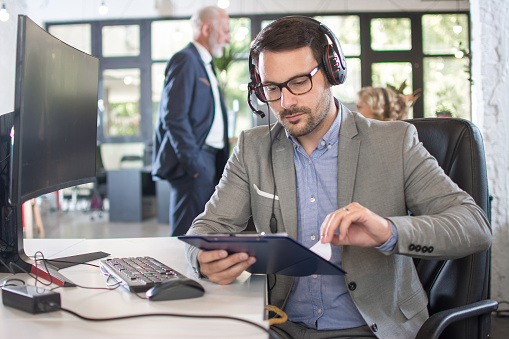 Concentrated young man in business wear talking on hands-free device and holding documents while sitting at his working place.