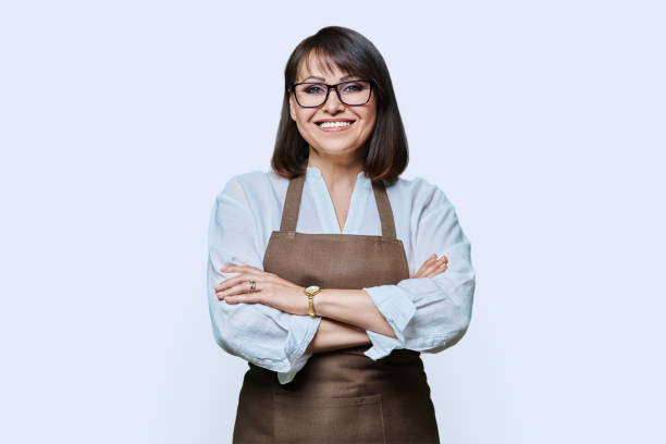 Confident middle aged woman in apron looking at camera on white background stock photo
