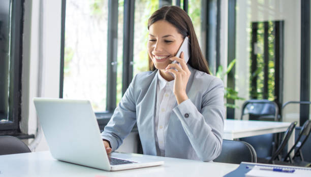 istock Smiling businesswoman talking on mobile phone and using laptop in office. 1446076971