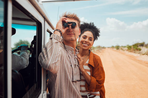 Road trip, travel and view with a couple using binoculars for sightseeing while outdoor on sand road during their summer vacation. Diversity, love and holiday with a man and woman traveling on a trip