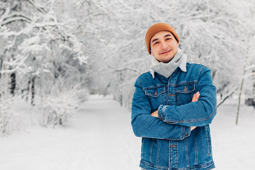 Portrait of stylish man in snowy winter park wearing jeans coat hat posing outdoors. Handsome looks at camera. Male fashion. Space