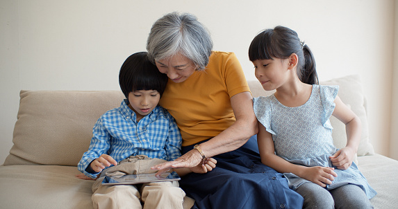 A grandmother with her two grandchildren are sitting on a couch in a domestic living room, playing with a digital tablet.