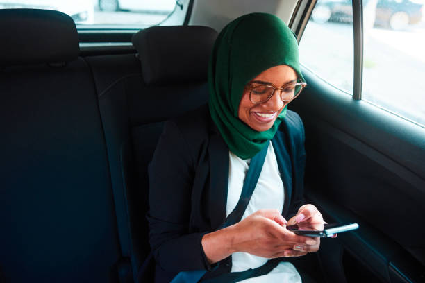 улыбающаяся мусульманская бизнесвумен переписывается по телефону во время езды в такси - mobile phone seat belt text messaging smiling стоковые фото и изображения