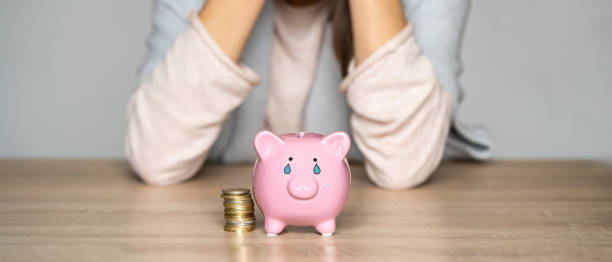 Sad girl with a crying piggy bank. Sad girl leaned on the table, covering her face with hands, in the foreground a crying piggy bank with painted tears. Woman lost her savings, was fired, and is experiencing financial difficulties. money cry stock pictures, royalty-free photos & images