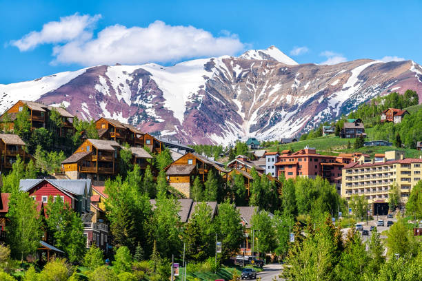 mount crested butte ski resort town, colorado with houses homes, wooden lodge hotels on hill in summer and green trees by snow-capped mountains in background - estância de esqui imagens e fotografias de stock