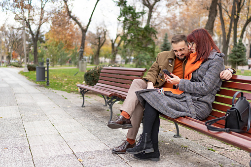 Happy young couple sitting together on bench in park using mobile phone