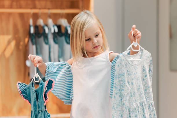 child, fashion and shopping while choosing kids clothing in a shop, store or boutique with a cute girl holding dresses thinking or picking color choice. little kid shopper or customer buying clothes - boutique shopping retail mother imagens e fotografias de stock