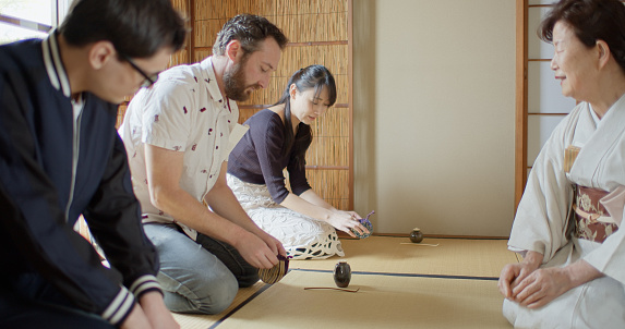 Two caucasian men and a Japanese woman attending a traditional Japanese tea ceremony at a Chashitsu in Tokyo, Japan, hosted by an elderly Japanese woman wearing a white kimono.