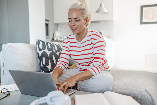 Photo of a mature woman sitting relaxed on the sofa, smiling and using a laptop