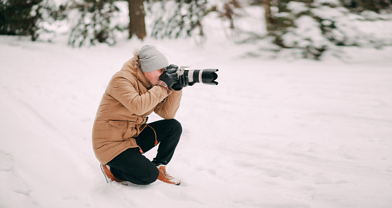 Man with camera in winter forest. Photographer takes pictures or shooting video of trees under snow, scenery. Nature photographer tourist with camera shoots. Leisure activity in cold season