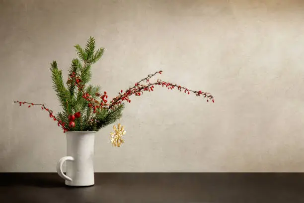 Still life with white ceramic cup and bouquet of spruce, barberry twigs and wild rose fruits on gray wall background, copy space