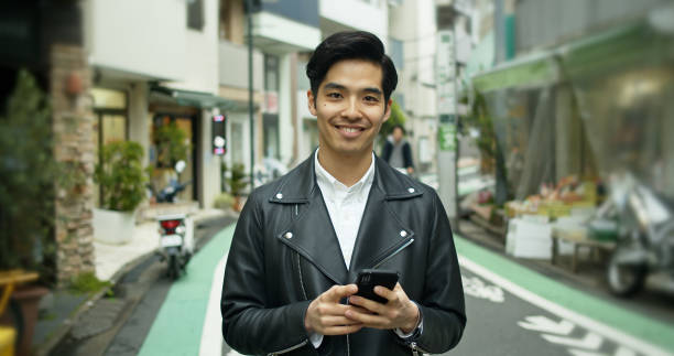istock Young Japanese Man Holding Cell Phone Smiling at Camera 1446111709