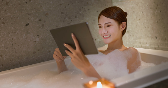 happy asian woman using tablet enjoy take a bubble foam bath inside bathtub in bathroom