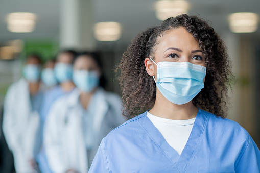 A nurse wearing blue scrubs smiles while a group of doctors and nurses stand behind her.