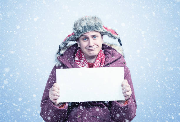 istock Pitiful frozen man in winter clothes holds an empty white poster in his hands, it is snowing on a blue background 1446122500