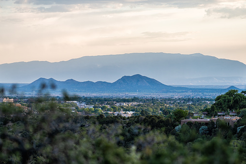 Colorful sunset in Santa Fe, New Mexico skyline with green foliage in foreground and cityscape buildings with mountains silhouette