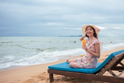Young Asian happy woman wearing sun hat holding orange juice relaxing on chair at the beach with happily.