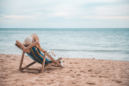 Hermosa joven asiática con sombrero brazo hacia arriba relajándose en la silla de playa, concepto de vacaciones felices de verano. photo