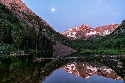Maroon Bells lake reflection with view of peaks in Aspen, Colorado at blue hour dawn in rocky mountains with snow in July summer and moon setting