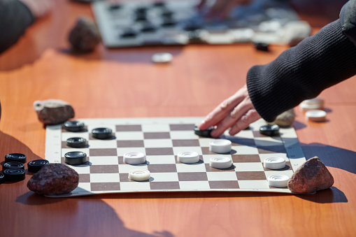 Outdoor checkers tournament on paper checkerboard on table, close up players hands. Outdoor draughts board game between two amateur players at sunny day, development of strategic thinking