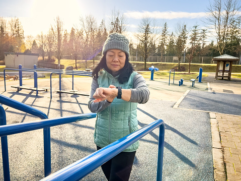 Chinese woman at a public outdoor park checking smartwatch while using exercise equipment.  North Vancouver, British Columbia, Canada.