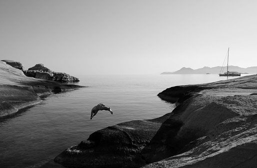 A grayscale shot of a man jumping into the water from the shore