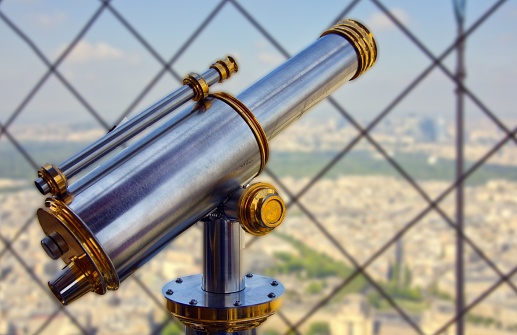 A closeup of shiny telescope by mesh wire fence with blur cityscape in the background