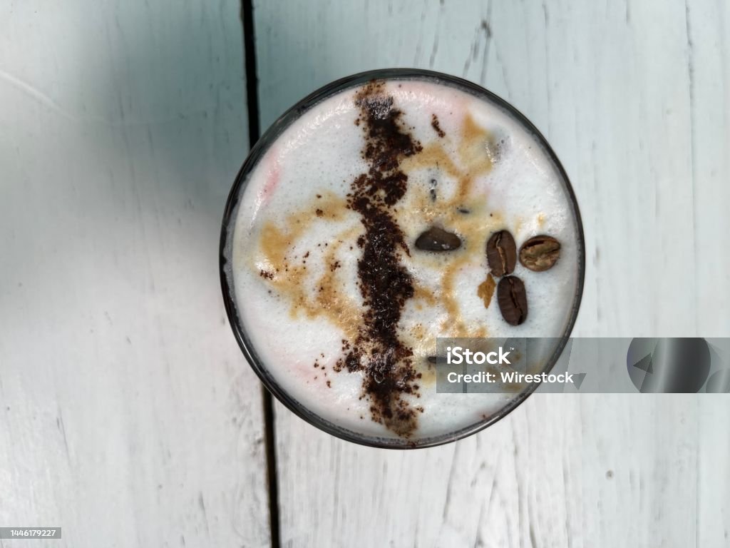 Top View of a Decorative Caffeinated Cappuccino Beverage Top View of a Decorative Caffeinated Cappuccino Beverage. Garnished with caramel, cacao, coffee bean, and foam.  Sitting on white wooden patio table Above Stock Photo