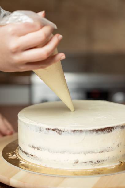 foto vertical de uma mulher decorando um bolo coberto com chantilly, com um saco de cachimbo, na cozinha - baking cake making women - fotografias e filmes do acervo