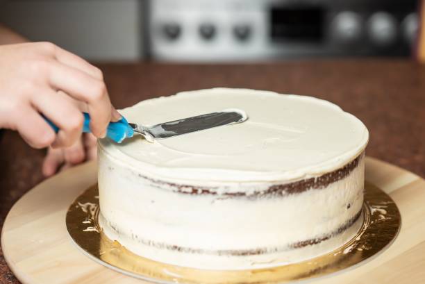 mulher alisando bordas de um bolo de chocolate coberto com chantilly, com uma espátula, na cozinha - baking cake making women - fotografias e filmes do acervo