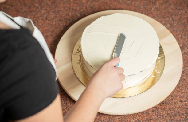 vista sobre um ombro de uma mulher alisando o topo do bolo com uma espátula, na cozinha - baking cake making women - fotografias e filmes do acervo