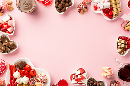 Valentine's Day concept. Top view photo of heart shaped saucers with sweets candies cookies and glass cups with beverage on isolated light pink background with blank space in the middle