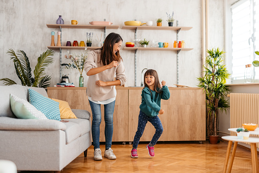 Young Asian mother and her multiracial daughter dancing together in the living room.