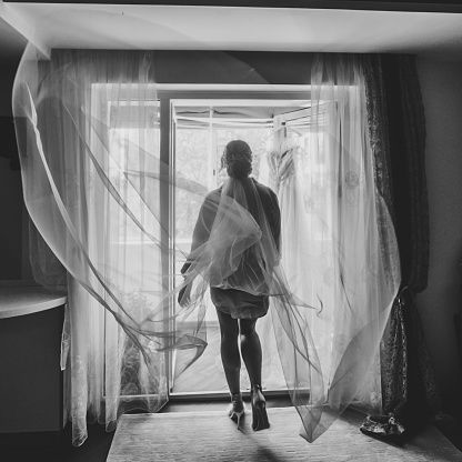 Veil. Selective focus. Portrait of a stylish bride with an elegant long veil near window. Beautiful bride style. Back view. Black and white photo.