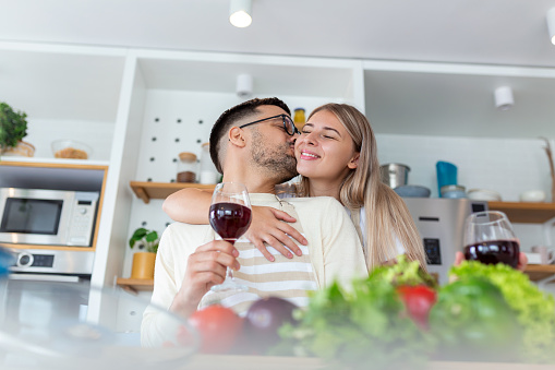 Affectionate young man kissing his wife while cooking together. Beautiful young couple is talking and smiling while cooking healthy food in kitchen at home.Man is kissing his girlfriend in cheek