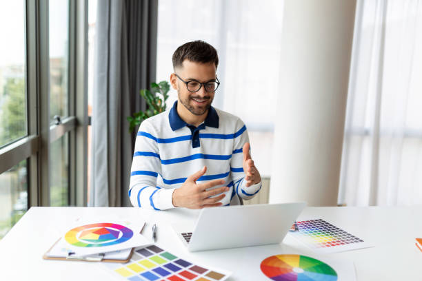istock Cheerful man designer having video conference with clients, sitting at desk in front of computer, holding color palettes, gesturing and smiling, copy space 1446204412