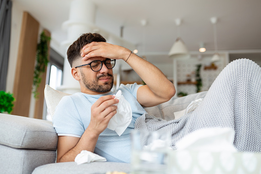 Man feeling sick lying in the bed and looking the thermometer. Sick man lying on sofa checking his temperature under a blanket at home in the living room