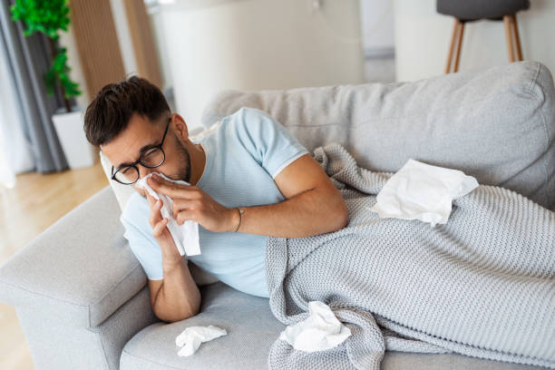 istock Sick man lying on sofa at home and blowing nose. Man wrapped in plaid lying on the sofa feeling sick illness at home 1446206538