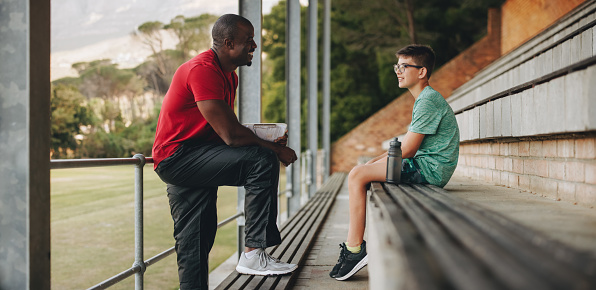 Sports coach talking to his student outside in a school. Physical education teacher guiding a young school boy. Mentoring and motivating an elementary school kid.