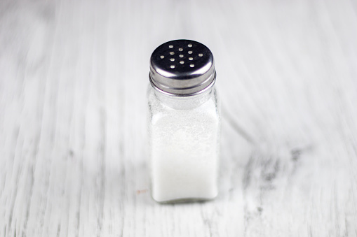 Glass salt shaker shot on rustic wooden table. A spoon with salt is beside the shaker placed directly on the table. The composition is at the left of an horizontal frame leaving useful copy space for text and/or logo at the right. Predominant colors are white and brown. High resolution 42Mp studio digital capture taken with Sony A7rII and Sony FE 90mm f2.8 macro G OSS lens