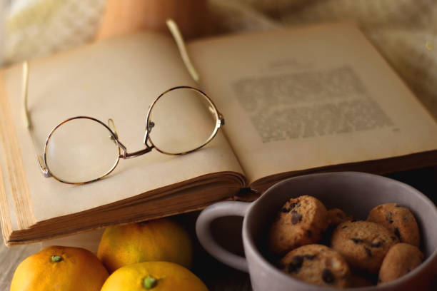 Cup of tea or coffee, seasonal spices, bowl of cookies, blanket, pumpkins, colorful leaves, books and tangerines on wooden table. Cozy hygge at home. Selective focus.