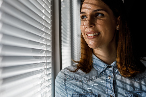 Copy space shot of happy young woman standing by the window, looking outside, smiling and pondering. Blinds casting interesting shade on her face.