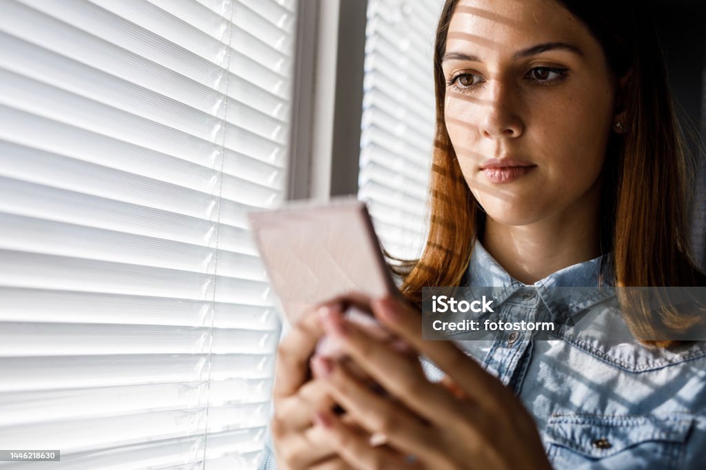 Young woman using smart phone by the window Portrait of happy young woman using smart phone by the window in her room. Blinds casting interesting shade on her face. Communication Stock Photo