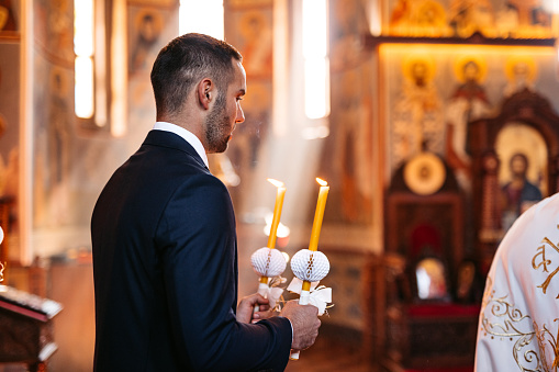 Handsome young groom having an orthodox church wedding ceremony. Holding lit up candles.