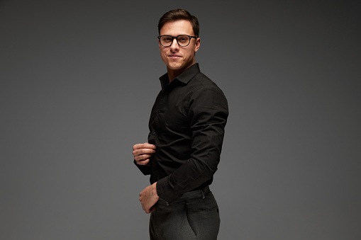 Looks happy. Confident young man in black business classic style shirt and grey trousers isolated over grey background. Fashion look of businessman. Concept of business, youth, modern lisfestyle.