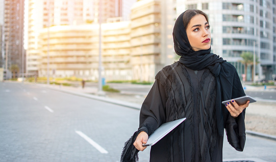 Beautiful young muslim woman with laptop and phone on the street.