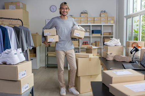 Confident young Asian man retail seller, entrepreneur, online store drop shipping small business owner looking at camera standing in delivery shipping warehouse with parcel boxes. Looking at camera.
