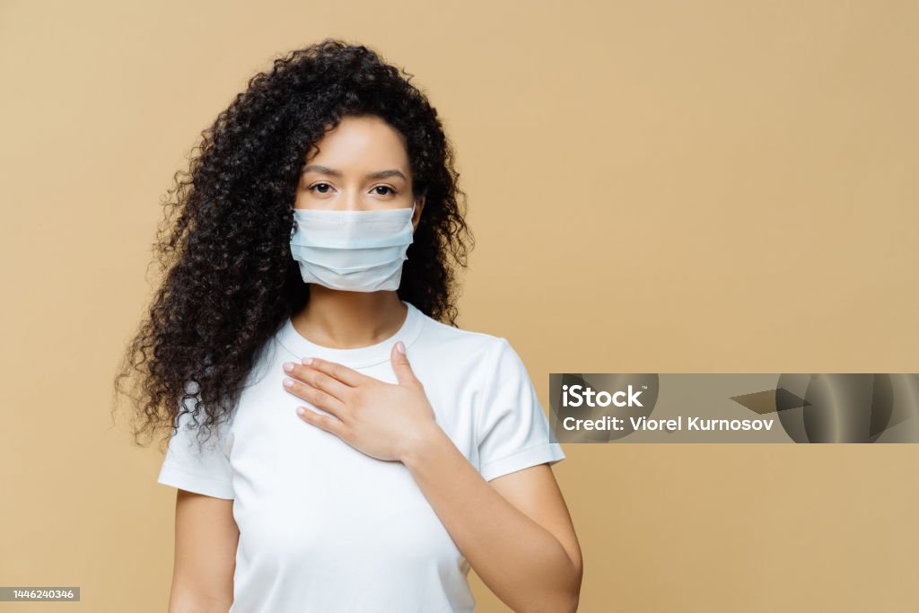 Serious Afro American woman wears medical face mask, has problems with breathing, presses hand to chest, got infected with coronavirus, isolated on beige ackground. Covid 19, health care concept 20-24 Years Stock Photo
