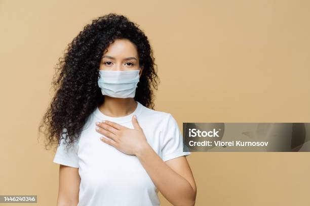 Serious Afro American Woman Wears Medical Face Mask Has Problems With Breathing Presses Hand To Chest Got Infected With Coronavirus Isolated On Beige Ackground Covid 19 Health Care Concept Stock Photo - Download Image Now