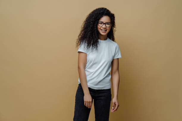 istock Smiling dark skinned young woman with glad face expression, wears optical glasses, white t shirt and jeans, poses carefree against beige background. People, happy mood and lifestyle concept. 1446240356